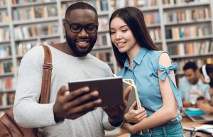 Two students in a library looking at a tablet and smiling.