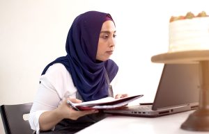 Woman wearing a hijab holding a notebook and looking at laptop