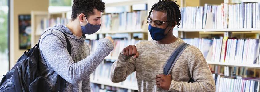 Two college students in library bump fists, face masks graduate silhouette 2019 2020